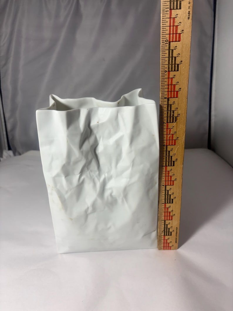 Puede incluir: Un florero de cer&aacute;mica blanca con forma de bolsa de papel arrugada, colocado junto a una regla de madera que muestra que el objeto mide aproximadamente 21,5 cm de altura. El florero tiene un acabado mate y texturas irregulares y plegadas.