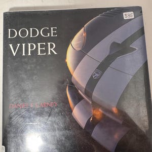 Op de afbeelding: Een boek getiteld "Dodge Viper" door Daniel E. Carney. De omslag toont een close-up van een zilveren en zwarte Dodge Viper auto met een witte streep. Het boek ligt op een witte ondergrond.