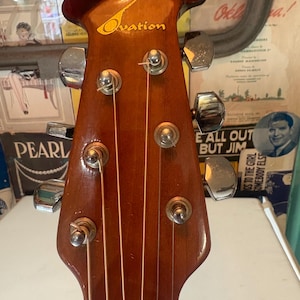 May include: Close-up of an Ovation acoustic guitar headstock. The headstock is made of dark brown wood with the brand name in gold lettering. Six chrome tuning pegs are visible, along with the strings. The guitar is in good condition.