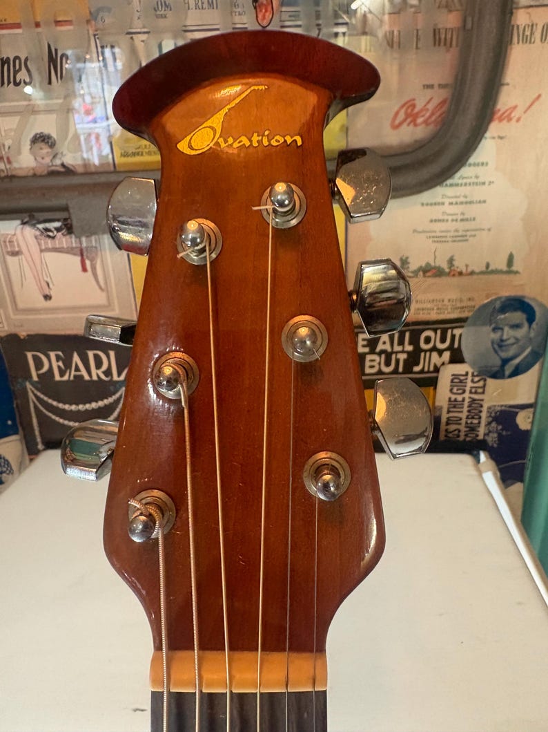 May include: Close-up of an Ovation acoustic guitar headstock. The headstock is made of dark brown wood with the brand name in gold lettering. Six chrome tuning pegs are visible, along with the strings. The guitar is in good condition.