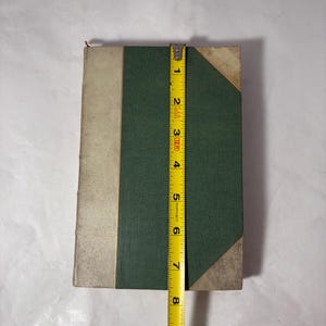 Puede incluir: Un libro antiguo con una cubierta verde y un lomo crema. El libro mide aproximadamente 20 cm de alto. Las esquinas del libro est&aacute;n reforzadas con un material claro. Una cinta m&eacute;trica amarilla se coloca junto al libro.