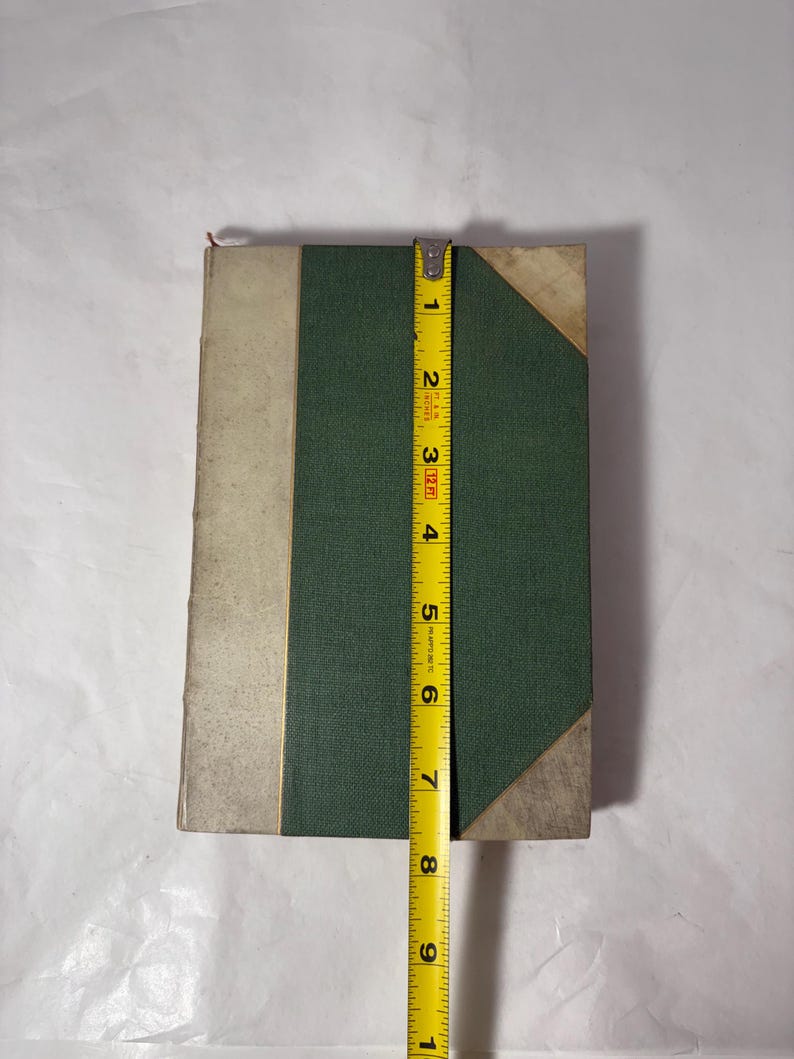 Puede incluir: Un libro antiguo con una cubierta verde y un lomo crema. El libro mide aproximadamente 20 cm de alto. Las esquinas del libro est&aacute;n reforzadas con un material claro. Una cinta m&eacute;trica amarilla se coloca junto al libro.