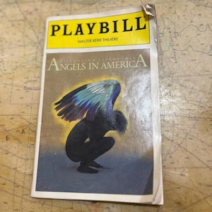 Op de afbeelding: Een vintage programmaboekje voor "Angels in America" in het Walter Kerr Theatre. De omslag toont een figuur met kleurrijke vleugels. De titel staat in vette gele letters. Het programmaboekje is licht versleten, met een gekreukelde hoek.