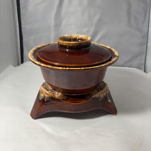 Op de afbeelding: Een bruine keramische ovenschaal met deksel, op een bijpassende standaard. De schaal en het deksel zijn effen, glanzend bruin, met een lichtere, getextureerde rand. De standaard heeft een vergelijkbaar bruin glazuur.