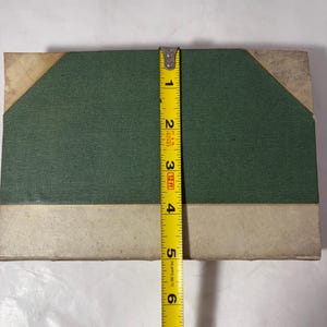 Puede incluir: Un libro rectangular con una cubierta de tela verde y detalles beige. El libro mide aproximadamente 15 cm de altura. Una cinta m&eacute;trica amarilla se coloca verticalmente a lo largo del borde del libro.