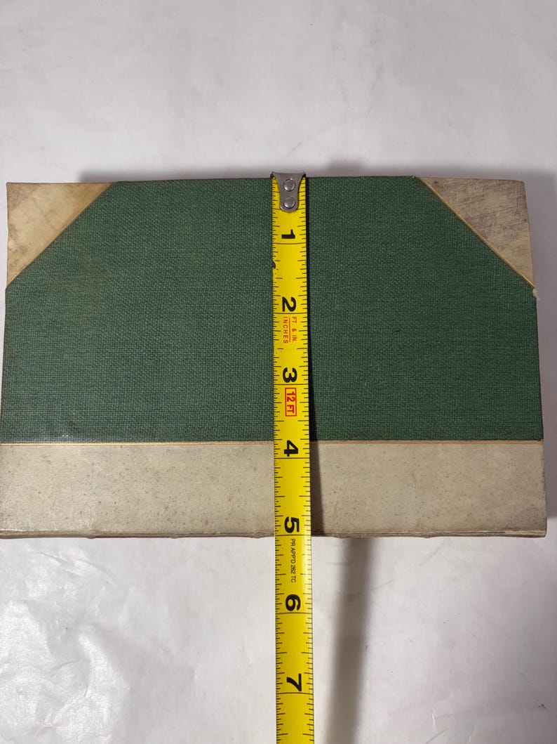 Puede incluir: Un libro rectangular con una cubierta de tela verde y detalles beige. El libro mide aproximadamente 15 cm de altura. Una cinta m&eacute;trica amarilla se coloca verticalmente a lo largo del borde del libro.