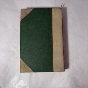 Puede incluir: Un libro antiguo con una cubierta verde texturizada y lomo beige. Las esquinas est&aacute;n reforzadas con material beige y acentuadas con ribetes dorados. Un marcador de hilo rojo sobresale de la parte superior.