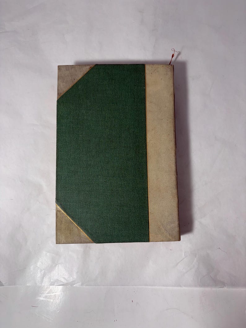Puede incluir: Un libro antiguo con una cubierta verde texturizada y lomo beige. Las esquinas est&aacute;n reforzadas con material beige y acentuadas con ribetes dorados. Un marcador de hilo rojo sobresale de la parte superior.
