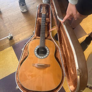 May include: An acoustic guitar with a light brown wooden body and a dark brown neck rests inside an open brown case. The guitar has a round sound hole with a decorative rosette and six strings. The case is lined with a soft, brown material.