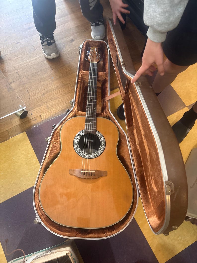 May include: An acoustic guitar with a light brown wooden body and a dark brown neck rests inside an open brown case. The guitar has a round sound hole with a decorative rosette and six strings. The case is lined with a soft, brown material.