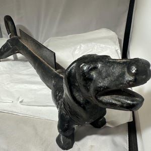 May include: A black cast iron dachshund dog-shaped doorstop. The dog has an open mouth and a slot on its back. The doorstop is designed to hold a door open and has a handle on the back.