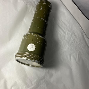 Könnte beinhalten: Ein olivgrünes zylindrisches Objekt mit strukturierter Oberfläche. Das Objekt hat eine kreisförmige Basis und eine etwas breitere Oberseite. Auf der Seite ist ein weißer Aufkleber mit der Nummer "1875" angebracht. Das Objekt scheint aus Metall zu sein.