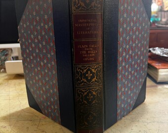 Op de afbeelding: Een vintage hardcover boek getiteld "Immortal Masterpieces of Literature" met "Plain Tales From The Hills Ballads" van Kipling. Het boek heeft een donkerbruine rug met gouden letters en sierlijke ontwerpen. De kaften zijn blauw met een rood-wit bloemmotief.