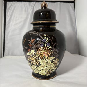 May include: A black ceramic ginger jar with a lid, featuring floral and dragonfly designs in yellow, orange, and purple. The jar has gold trim and a gold finial on the lid. The overall design is ornate and decorative.
