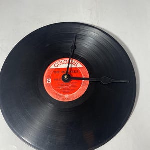 May include: A black vinyl record clock with a red and white label featuring the text "The Monkees". The clock hands are black and point to the time. The clock is a unique decorative item.