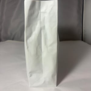 Puede incluir: Una bolsa blanca alta y rectangular con un lado reforzado y fondo plano. La bolsa est&aacute; hecha de un material mate y opaco y presenta pliegues verticales a lo largo del centro y los lados, lo que indica una estructura flexible de papel o pl&aacute;stico.