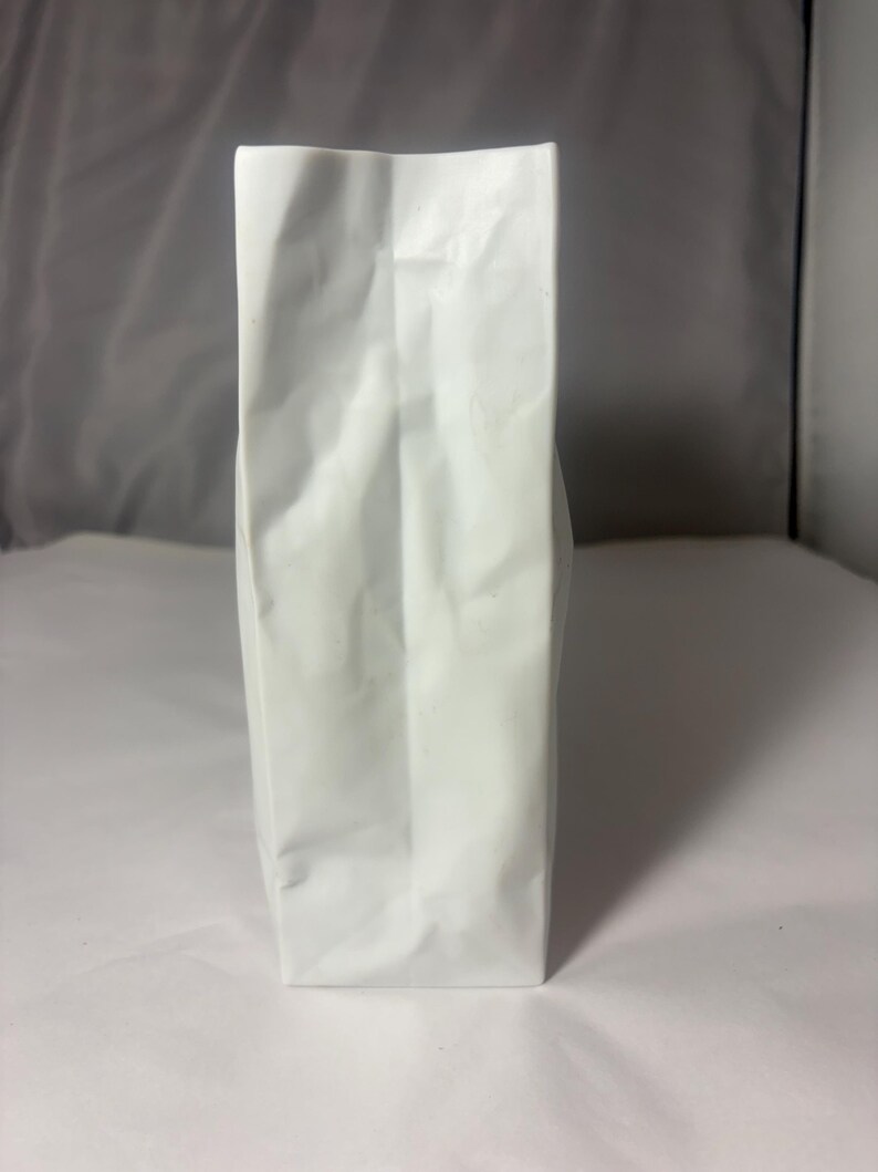 Puede incluir: Una bolsa blanca alta y rectangular con un lado reforzado y fondo plano. La bolsa est&aacute; hecha de un material mate y opaco y presenta pliegues verticales a lo largo del centro y los lados, lo que indica una estructura flexible de papel o pl&aacute;stico.
