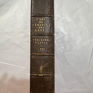 May include: A dark brown leather-bound book spine with gold embossed lettering that reads L'ART DE VERIFIER LES DATES, TROISIEME PARTIE, and XIII. The spine features five raised horizontal bands and a textured, aged finish with visible wear.