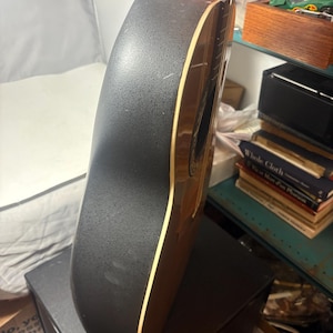 May include: Acoustic-electric guitar with a black textured back and sides. The guitar has a brown wooden neck and soundhole, with a cream-colored binding. The instrument is resting on a black surface.