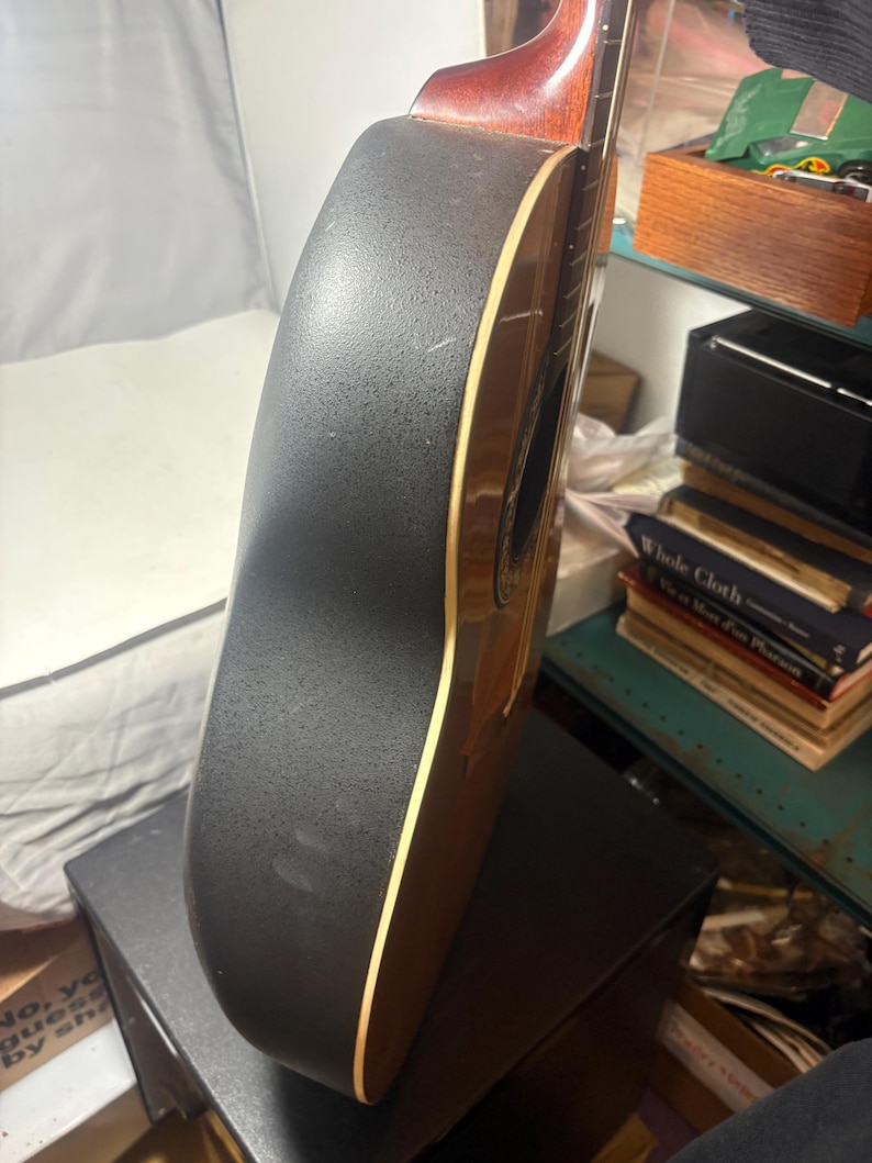 May include: Acoustic-electric guitar with a black textured back and sides. The guitar has a brown wooden neck and soundhole, with a cream-colored binding. The instrument is resting on a black surface.