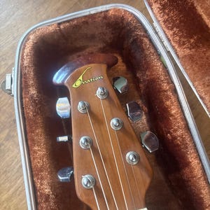 May include: Close-up of an acoustic guitar headstock inside a brown case. The headstock is made of wood and features the brand name "Ovation" in a gold script. The guitar strings are visible, along with the tuning pegs.