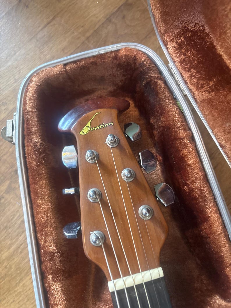 May include: Close-up of an acoustic guitar headstock inside a brown case. The headstock is made of wood and features the brand name "Ovation" in a gold script. The guitar strings are visible, along with the tuning pegs.