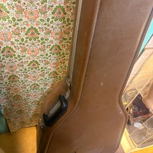 May include: A brown guitar case with a black handle and silver hardware. The case has a textured surface and the word "ovation" is printed on the side. The case is leaning against a floral patterned wall.