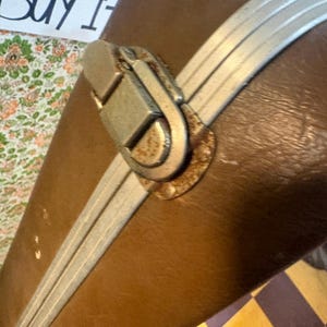 May include: Close-up of a vintage brown suitcase with silver-colored trim and a metal clasp. The suitcase has a worn appearance, suggesting age and use. The text "Buy It" is visible in the background.
