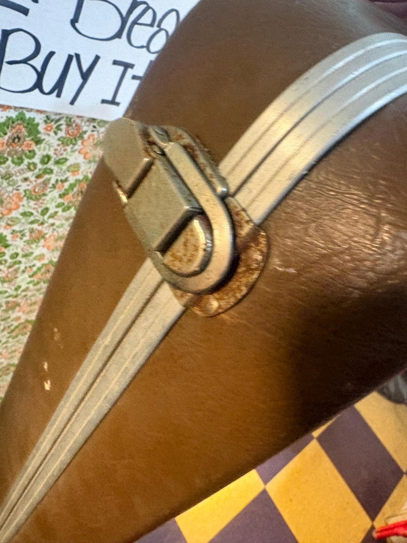 May include: Close-up of a vintage brown suitcase with silver-colored trim and a metal clasp. The suitcase has a worn appearance, suggesting age and use. The text "Buy It" is visible in the background.