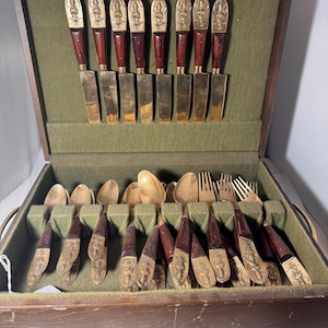 May include: A flatware set in a wooden storage case with a green felt interior. The set includes knives, spoons, and forks with dark wood handles and gold-toned metal accents featuring embossed figures of deities on the handle ends.
