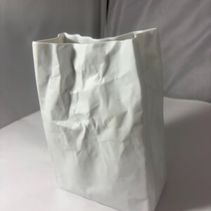 Puede incluir: Un florero de cer&aacute;mica blanca dise&ntilde;ado para parecerse a una bolsa de papel arrugada. El objeto presenta una base rectangular y un borde superior irregular y plegado con pliegues profundos en toda su superficie, imitando la textura del papel.