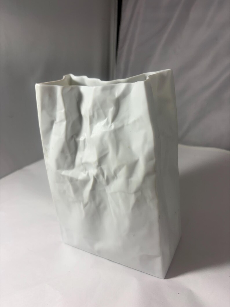 Puede incluir: Un florero de cer&aacute;mica blanca dise&ntilde;ado para parecerse a una bolsa de papel arrugada. El objeto presenta una base rectangular y un borde superior irregular y plegado con pliegues profundos en toda su superficie, imitando la textura del papel.