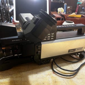 May include: A vintage RCA Newvicon video camera, with a black body and a beige and silver front panel. The camera has a prominent viewfinder and lens, with "AUTO FOCUS" and other controls visible. A black power cord is connected.