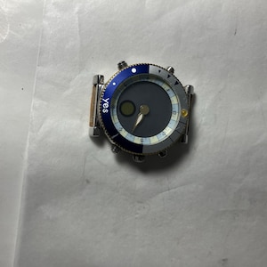 May include: A silver wristwatch with a blue and white bezel and a dark gray face. The watch has a round face with silver hands and a small green circle. The word "yes" is printed on the side of the watch.