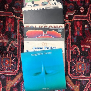May include: A collection of vinyl records with visible album art. The top record is labelled "DB Cooper." Other records include "Folk Song" and "tangerine dream." The records are on a patterned rug.
