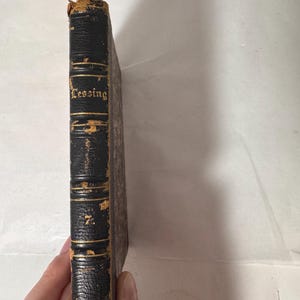 Könnte beinhalten: Ein altes Buch mit einem abgenutzten, schwarzen Lederrücken. Der Titel "Lessing" ist in goldener Schrift geprägt. Die Kanten des Buches weisen Alters- und Gebrauchsspuren auf. Das Buch wird von einer Hand gehalten. Es scheint eine Vintage-Ausgabe zu sein.