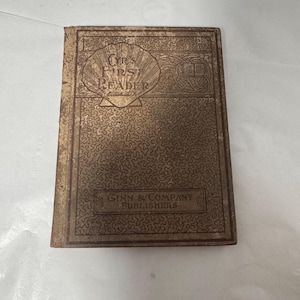 以下が含まれることがあります： Ginn & Company Publishersの「Our First Reader」というタイトルのヴィンテージ本。表紙は色あせた金色で、複雑なエンボス加工のデザインとテキストが施されています。本は使い込まれたようで、経年劣化の兆候が見られます。