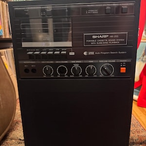 Può includere: Un sistema audio a cassetta portatile Sharp HK-25S nero con riproduzione slide/sync. Il dispositivo è dotato di un lettore di cassette, ingressi per microfono, manopole per volume e tono e un sistema di ricerca automatica dei programmi.