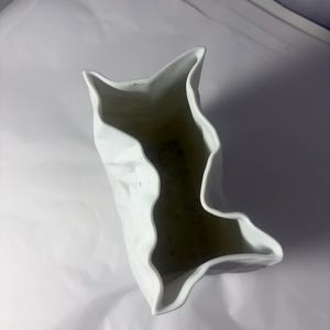 Puede incluir: Un florero de cer&aacute;mica blanca con una textura irregular, arrugada y plegada similar al papel. La abertura del recipiente presenta un borde dentado y ondulado que imita la apariencia de una bolsa arrugada, creando una forma escultural.