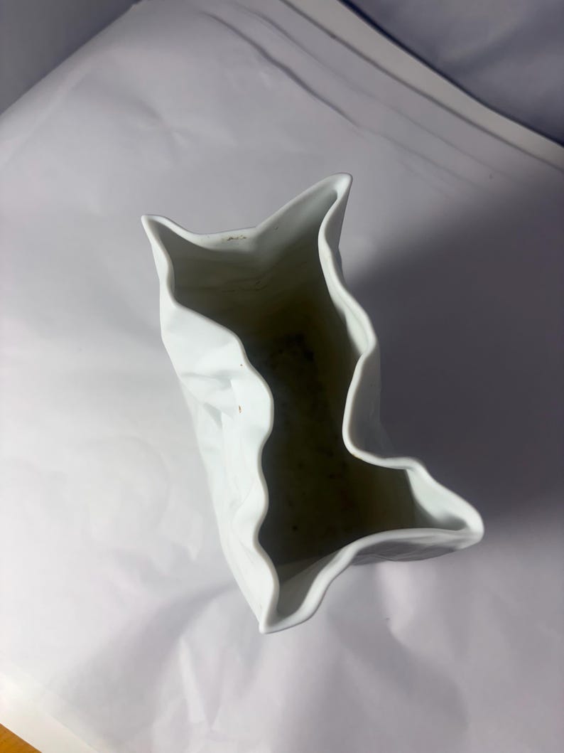 Puede incluir: Un florero de cer&aacute;mica blanca con una textura irregular, arrugada y plegada similar al papel. La abertura del recipiente presenta un borde dentado y ondulado que imita la apariencia de una bolsa arrugada, creando una forma escultural.