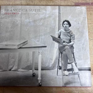 May include: A black and white photograph of a book cover with the title "FRANCISCA SUTIL" and the word "ALQUIMIA". The image shows a woman seated on a stool, holding a book. A table with a rectangular object is to the left.
