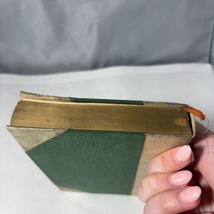Puede incluir: Un libro antiguo con una cubierta verde y bordes dorados. El lomo del libro es verde y las esquinas est&aacute;n reforzadas con metal plateado. Las p&aacute;ginas est&aacute;n envejecidas y un peque&ntilde;o hilo naranja est&aacute; unido al borde superior.