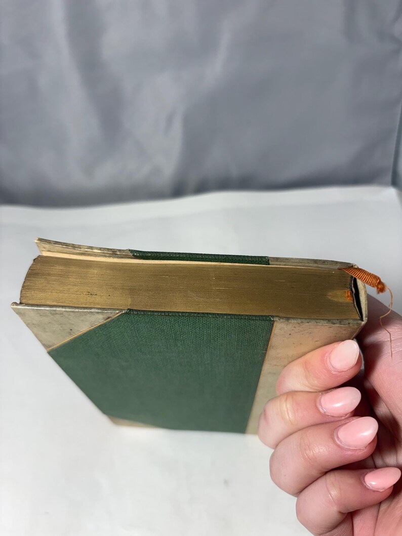 Puede incluir: Un libro antiguo con una cubierta verde y bordes dorados. El lomo del libro es verde y las esquinas est&aacute;n reforzadas con metal plateado. Las p&aacute;ginas est&aacute;n envejecidas y un peque&ntilde;o hilo naranja est&aacute; unido al borde superior.