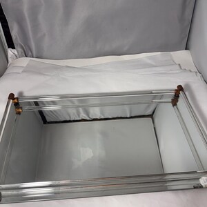 Könnte beinhalten: Ein rechteckiges Spiegeltablett mit einer reflektierenden Glasbasis und Seitenwänden. Das Tablett ist von klaren Glasscheiben umrahmt, die an den vier oberen Ecken durch kleine, dunkelbraune zylindrische Verbindungsstücke zusammengehalten werden.