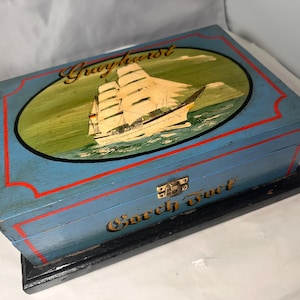 Puede incluir: Una caja de madera vintage pintada de azul claro con un borde rojo. La tapa presenta una pintura ovalada de un velero con el nombre "Grayhurst" encima. La caja tiene las palabras "Sweet Fuel" en la parte delantera.