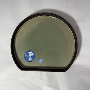 Könnte beinhalten: Ein runder, salbeigrüner Keramikteller mit einem dunkelbraunen Rand. Zwei kleine, blau-weiße kreisförmige Designs befinden sich auf der Oberfläche des Tellers. Der Teller hat eine einzigartige, halbkreisförmige Form, ideal zum Servieren von Vorspeisen oder kleinen Gerichten.