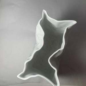 Puede incluir: Un jarr&oacute;n de cer&aacute;mica blanca con un borde distintivamente irregular, ondulado y plegado que crea una abertura en forma de estrella de m&uacute;ltiples puntas. El interior del recipiente es hueco y liso, mostrando un acabado blanco limpio y brillante.