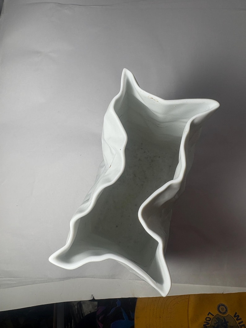 Puede incluir: Un jarr&oacute;n de cer&aacute;mica blanca con un borde distintivamente irregular, ondulado y plegado que crea una abertura en forma de estrella de m&uacute;ltiples puntas. El interior del recipiente es hueco y liso, mostrando un acabado blanco limpio y brillante.