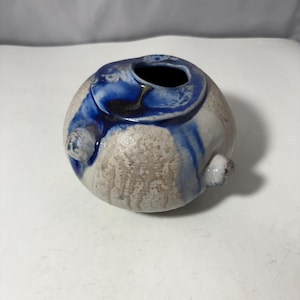 Op de afbeelding: Een kleine, ronde keramische vaas met een getextureerde, gebroken witte buitenkant en een donkerblauwe binnenkant. De vaas heeft een donkerblauw wervelontwerp en twee kleine, afgeronde uitsteeksels. De opening is cirkelvormig.