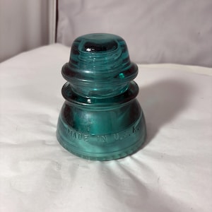 May include: A teal-colored glass electrical insulator with a tiered, rounded shape. The object features a smooth, translucent surface and embossed text along the base that reads MADE IN U.S.A. The design consists of three distinct stacked sections.